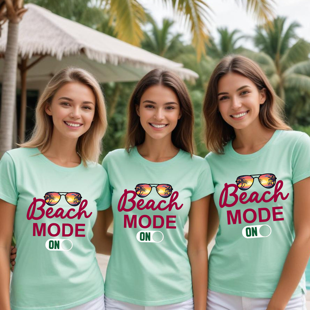 Three women wearing 'Beach Mode ON' t-shirts with sunglasses graphics in a tropical setting.
