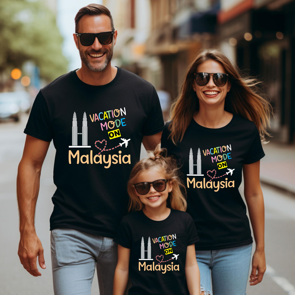 Family of three wearing matching black t-shirts with 'Vacation Mode ON Malaysia' text on a city street.